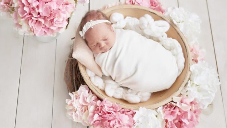 Bebê de faixa branca enrolado em lençol branco dentro de cesto ao redor de arranjos de flores
