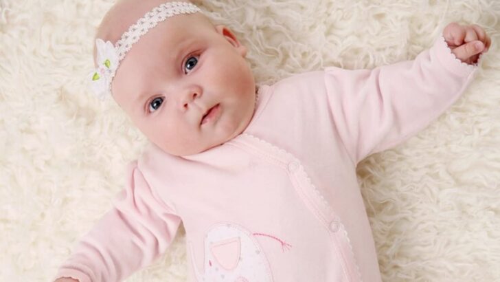 Bebê de faixa na cabeça e macacão rosa deitada em fundo fofo olhando para a câmera