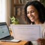 Mulher brasileira sorrindo em casa segurando um certificado impresso com texto borrado e um laptop com a plataforma Aprenda Mais MEC na tela também borrada